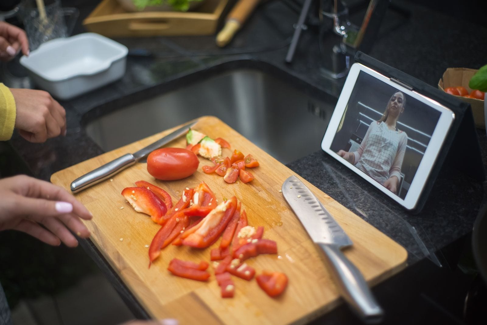 Hands preparing fresh vegetables on a chopping board beside a tablet showing a cooking video
